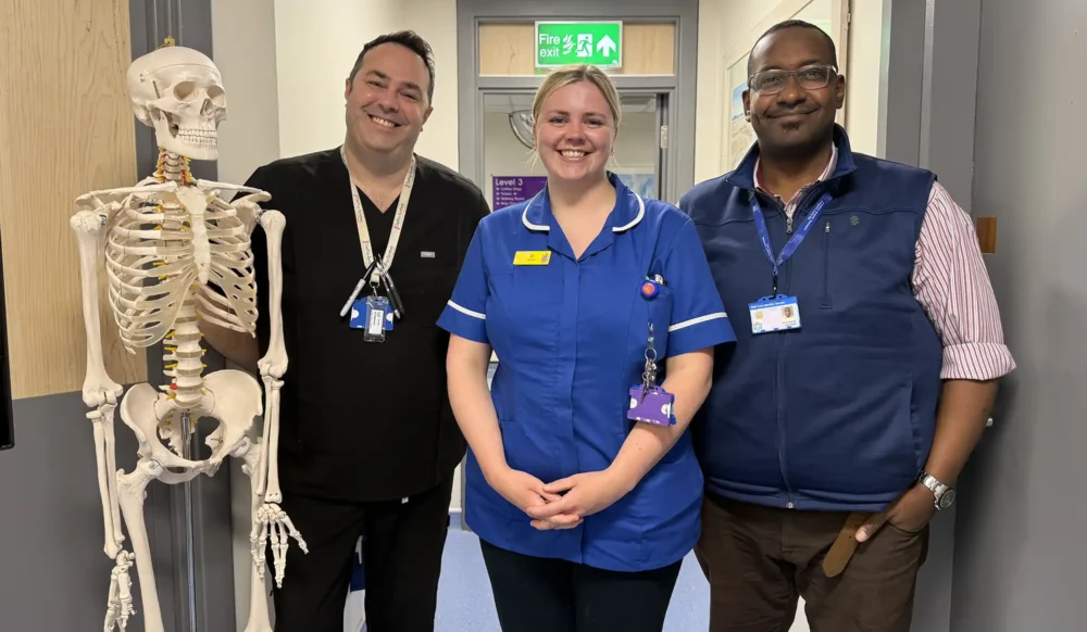 Three NHS staff from Yeovil spinal clinic stand in a row, with a skeleton model to their left, in what appears to be a ward at a hospital