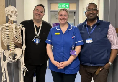 Three NHS staff from Yeovil spinal clinic stand in a row, with a skeleton model to their left, in what appears to be a ward at a hospital