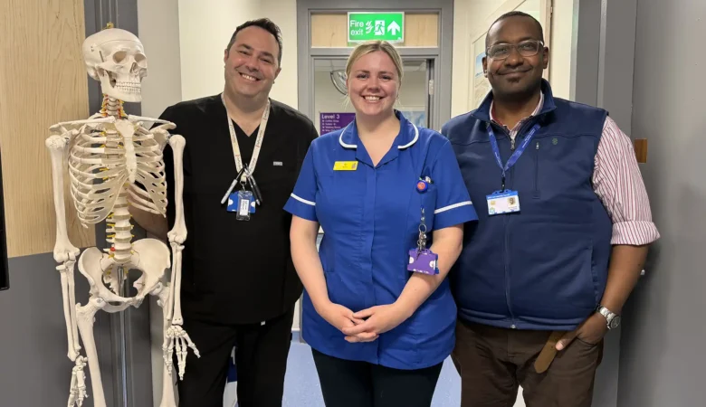 Three NHS staff from Yeovil spinal clinic stand in a row, with a skeleton model to their left, in what appears to be a ward at a hospital